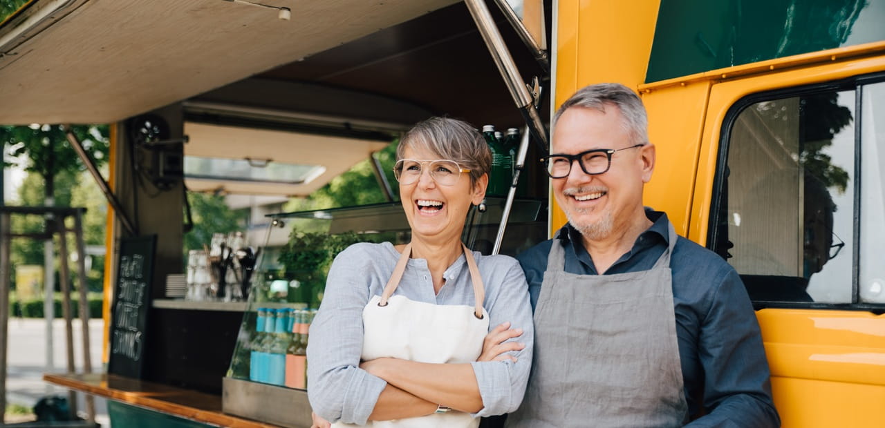 Retired couple in aprons smiling beside their food truck, starting a small business together in retirement.